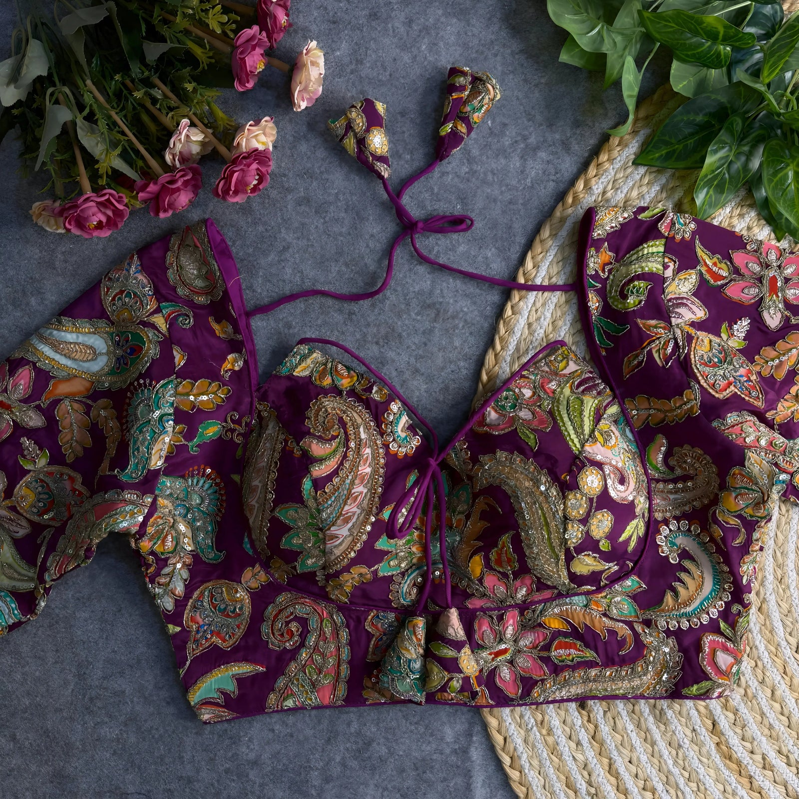 Purple padded blouse with intricate multicolor floral and paisley embroidery, featuring sequin accents and a tie-back detail, displayed on a grey surface with artificial pink flowers and green leaves in the background.