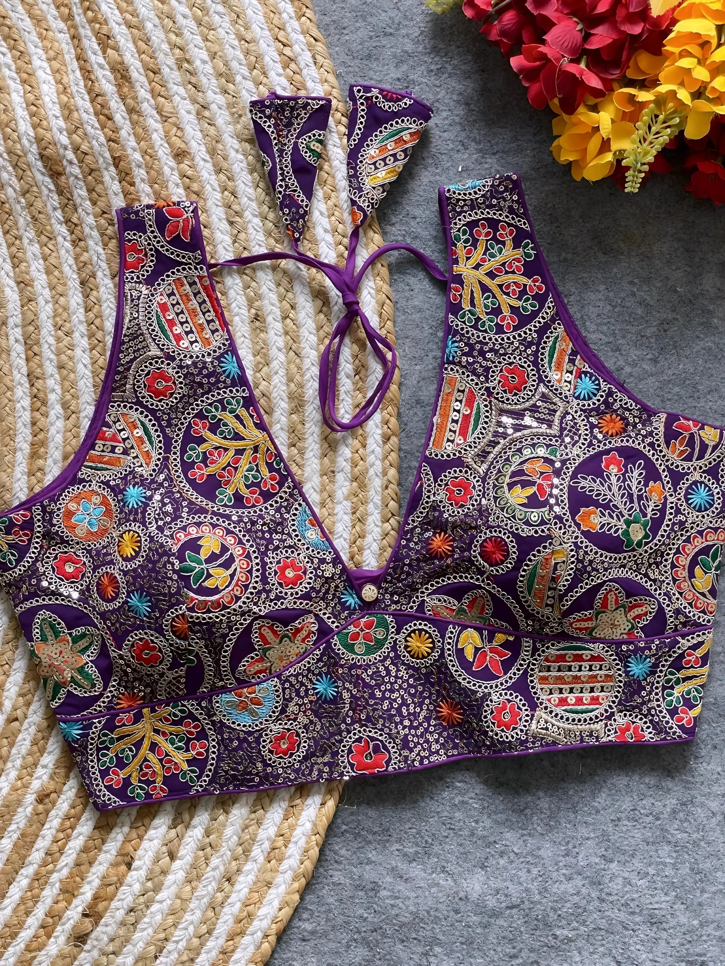 Traditional purple blouse featuring vibrant Madhubani-inspired multicolor embroidery with circular floral and geometric motifs, deep V-neck, and tie-back detail, displayed flat on a textured mat with artificial red and yellow flowers in the corner.