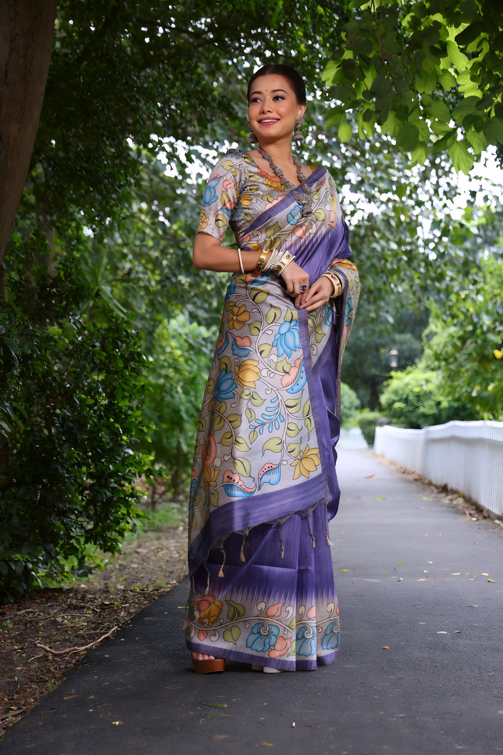 A woman models a stunning Lavender Purple Soft Tussar Silk Saree. The pallu features a wide, contrasting light grey Kalamkari-style print with large floral motifs in pale pink, blue, and yellow. She wears a matching short-sleeve printed blouse and accessorizes with heavy oxidized silver jewelry, posing outdoors in a garden.