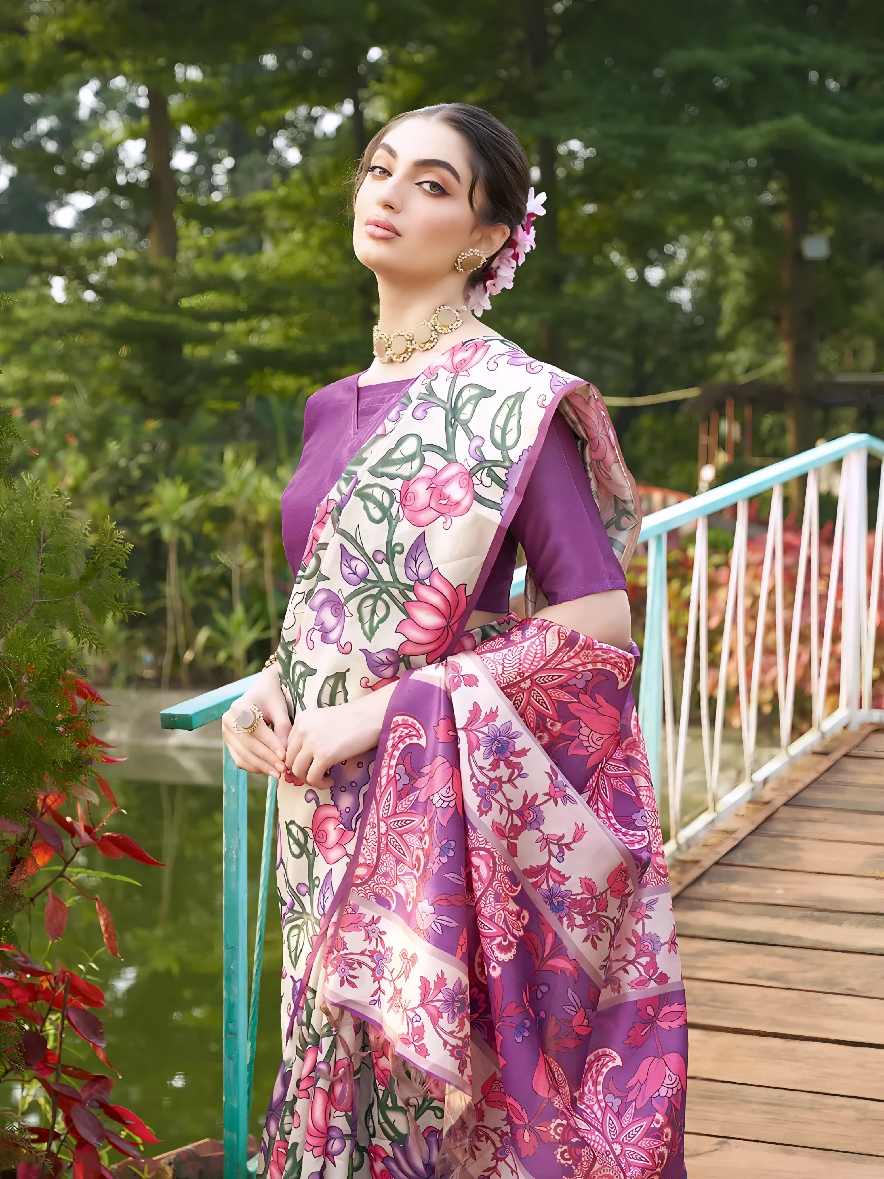 A woman models an elegant Beige Soft Tussar Silk Saree with a dense all-over print of purple, pink, and green floral and vine motifs. The pallu features a wide, contrasting deep purple panel with traditional weaving/print. She wears a matching short-sleeve purple blouse and a gold Kundan choker necklace, posing outdoors on a wooden footbridge.
