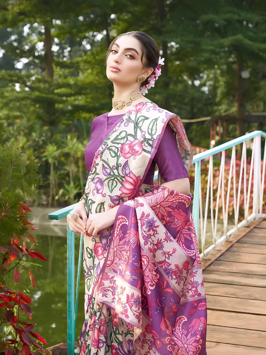 A woman models an elegant Beige Soft Tussar Silk Saree with a dense all-over print of purple, pink, and green floral and vine motifs. The pallu features a wide, contrasting deep purple panel with traditional weaving/print. She wears a matching short-sleeve purple blouse and a gold Kundan choker necklace, posing outdoors on a wooden footbridge.
