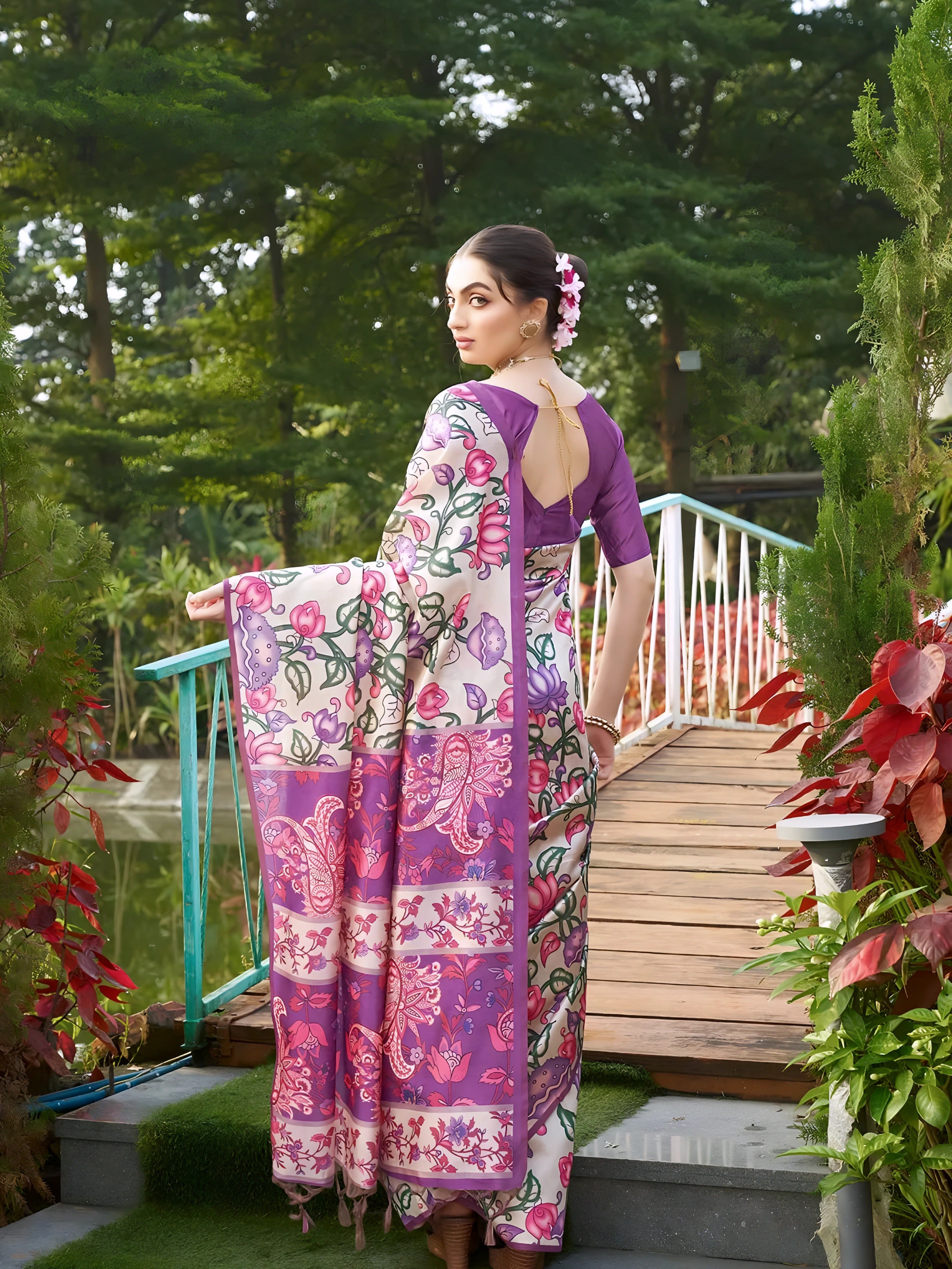 A woman models an elegant Beige Soft Tussar Silk Saree with a dense all-over print of purple, pink, and green floral and vine motifs. The pallu features a wide, contrasting deep purple panel with traditional weaving/print. She wears a matching short-sleeve purple blouse and a gold Kundan choker necklace, posing outdoors on a wooden footbridge.