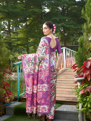 A woman models an elegant Beige Soft Tussar Silk Saree with a dense all-over print of purple, pink, and green floral and vine motifs. The pallu features a wide, contrasting deep purple panel with traditional weaving/print. She wears a matching short-sleeve purple blouse and a gold Kundan choker necklace, posing outdoors on a wooden footbridge.