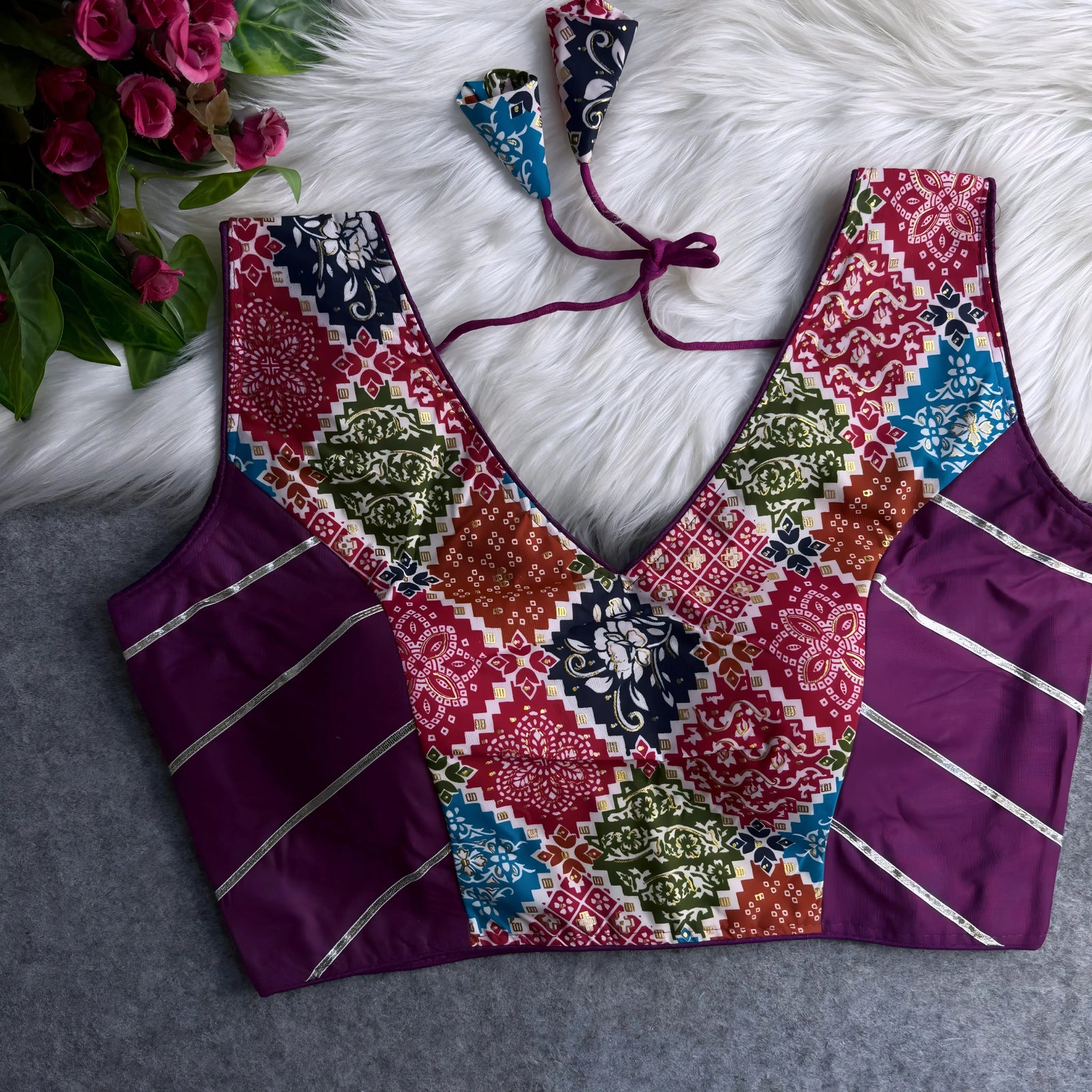 Traditional blouse with a vibrant multicolor Ajrak-inspired block print on the front panel, deep V-neckline, and rich purple silk side panels adorned with metallic silver stripes. Features a tasseled tie-back detail and is displayed flat on a white fur background with a bunch of pink artificial flowers in the corner.