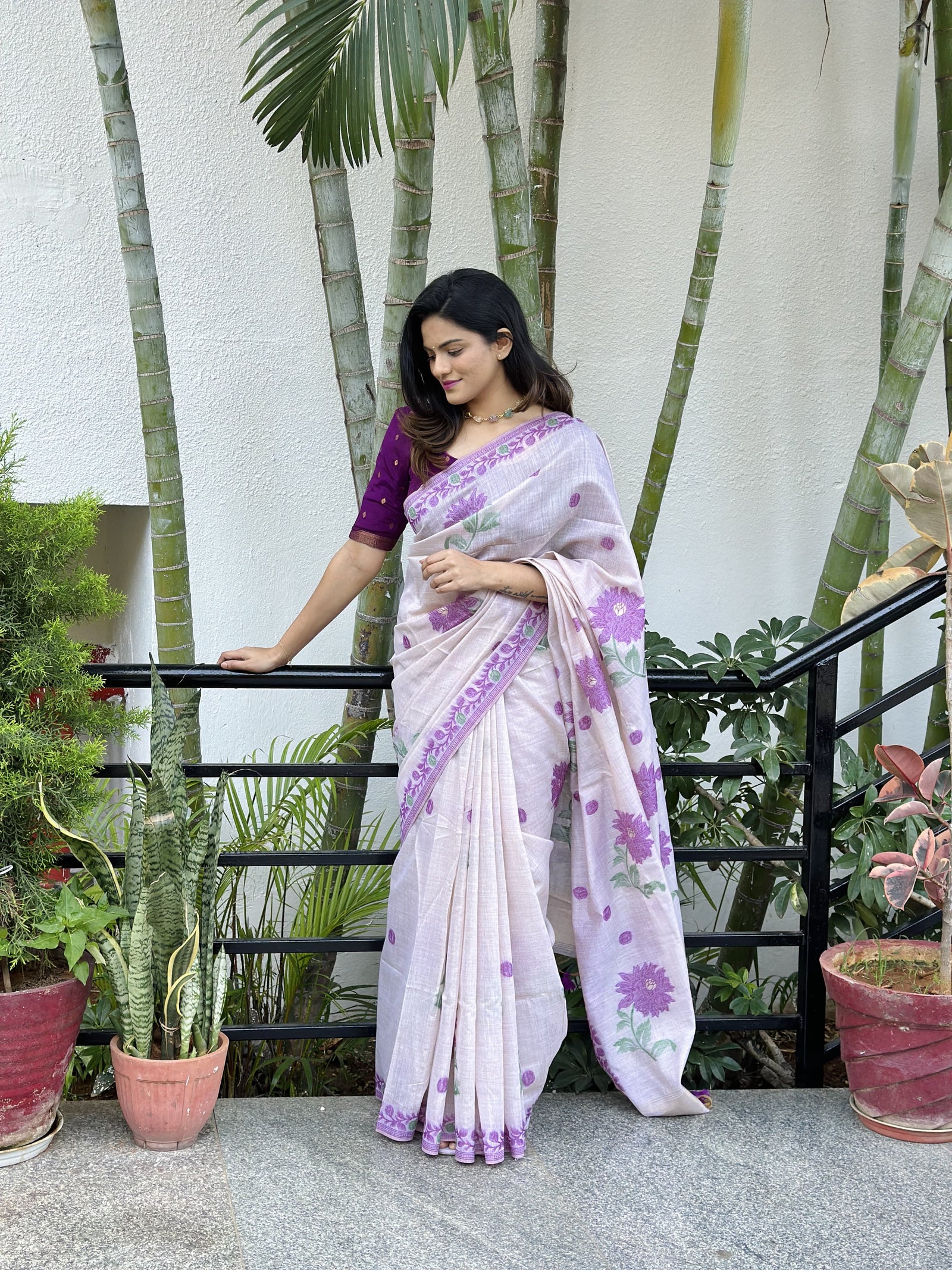 Woman in elegant purple resham weaving saree with floral motifs standing near plants and railing