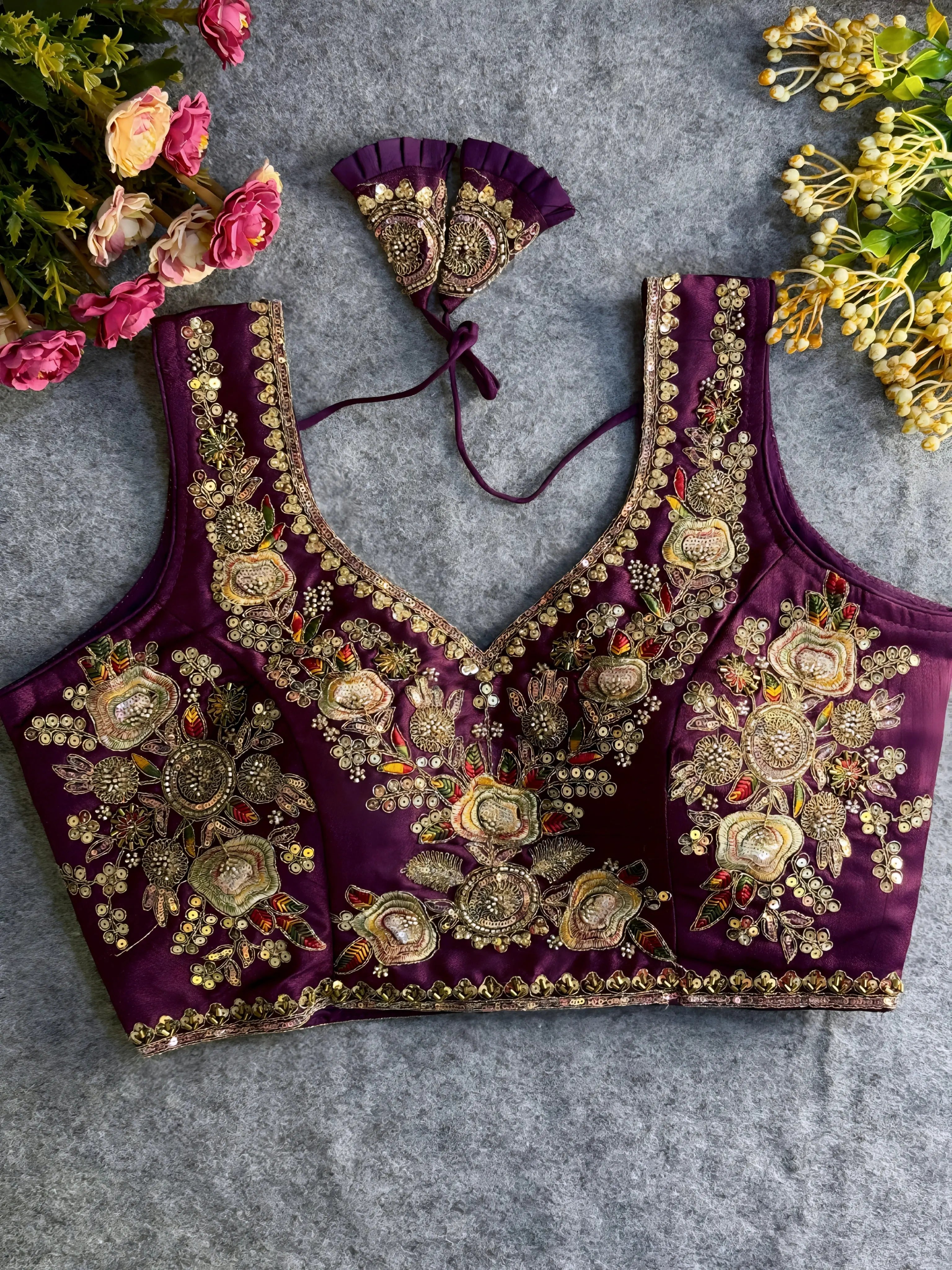 Traditional purple silk blouse featuring intricate gold zardozi embroidery and beadwork with floral and leaf motifs on the front and neckline, finished with decorative tassel ties, displayed flat on a textured grey background with artificial pink and yellow flowers in the corners.