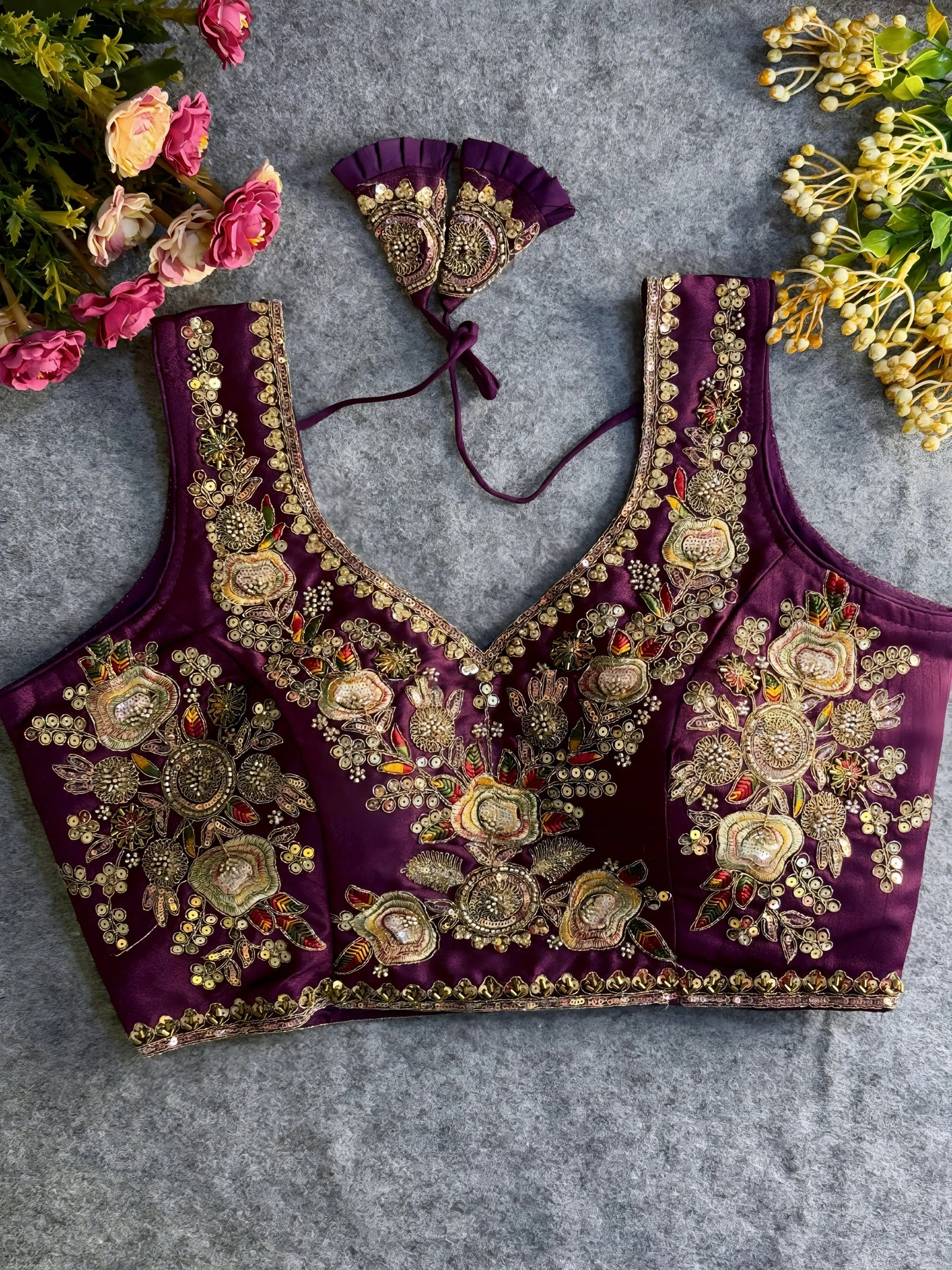 Traditional purple silk blouse featuring intricate gold zardozi embroidery and beadwork with floral and leaf motifs on the front and neckline, finished with decorative tassel ties, displayed flat on a textured grey background with artificial pink and yellow flowers in the corners.