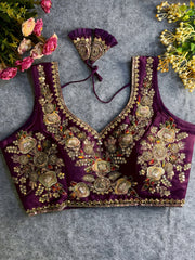 Traditional purple silk blouse featuring intricate gold zardozi embroidery and beadwork with floral and leaf motifs on the front and neckline, finished with decorative tassel ties, displayed flat on a textured grey background with artificial pink and yellow flowers in the corners.