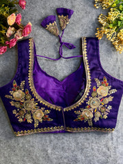 Traditional purple silk blouse featuring intricate gold zardozi embroidery and beadwork with floral and leaf motifs on the front and neckline, finished with decorative tassel ties, displayed flat on a textured grey background with artificial pink and yellow flowers in the corners.