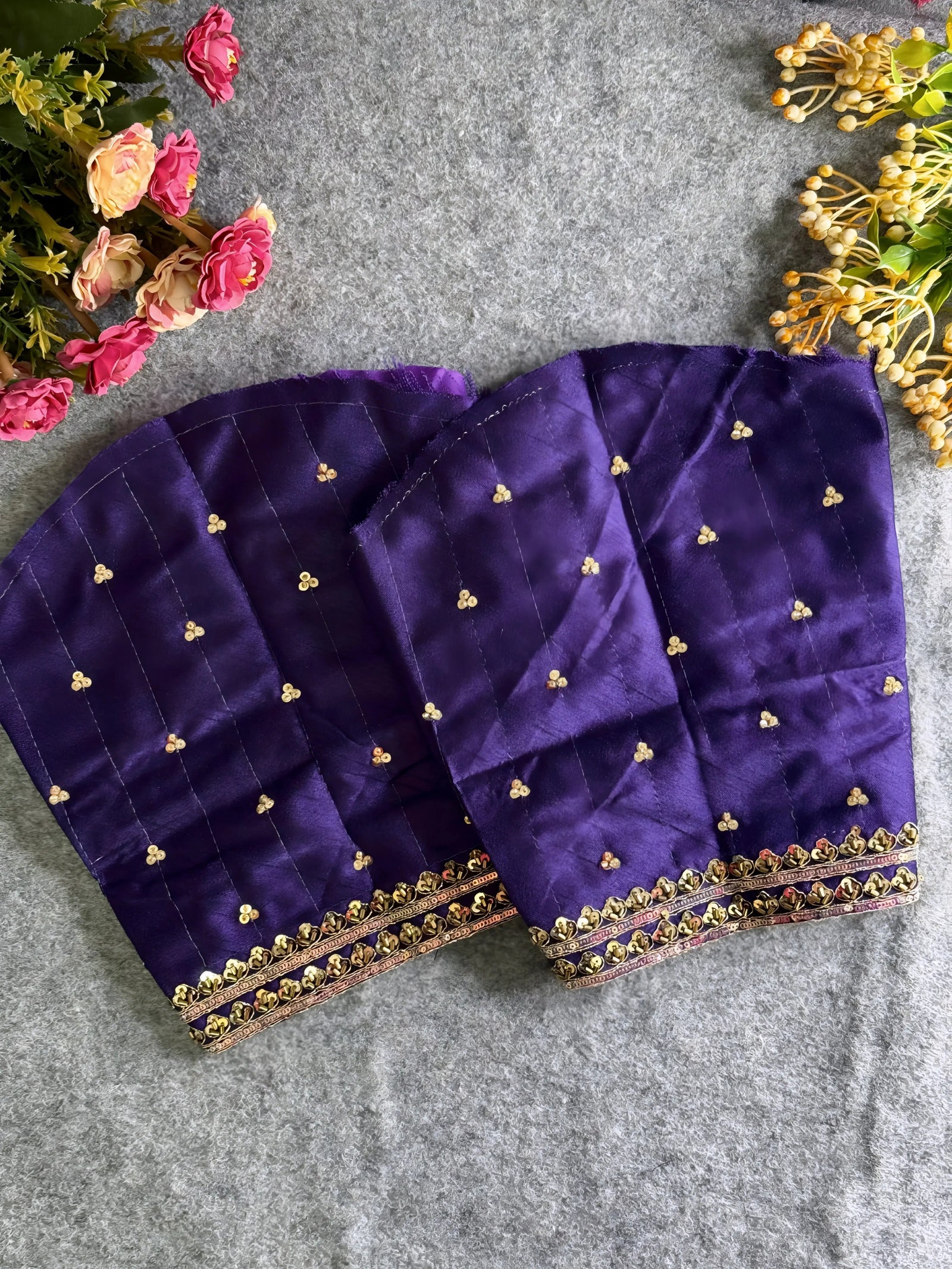 Traditional purple silk blouse featuring intricate gold zardozi embroidery and beadwork with floral and leaf motifs on the front and neckline, finished with decorative tassel ties, displayed flat on a textured grey background with artificial pink and yellow flowers in the corners.