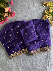 Traditional purple silk blouse featuring intricate gold zardozi embroidery and beadwork with floral and leaf motifs on the front and neckline, finished with decorative tassel ties, displayed flat on a textured grey background with artificial pink and yellow flowers in the corners.