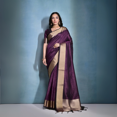 Woman wearing a purple silk saree with a gold border and traditional jewelry, standing against a blue background