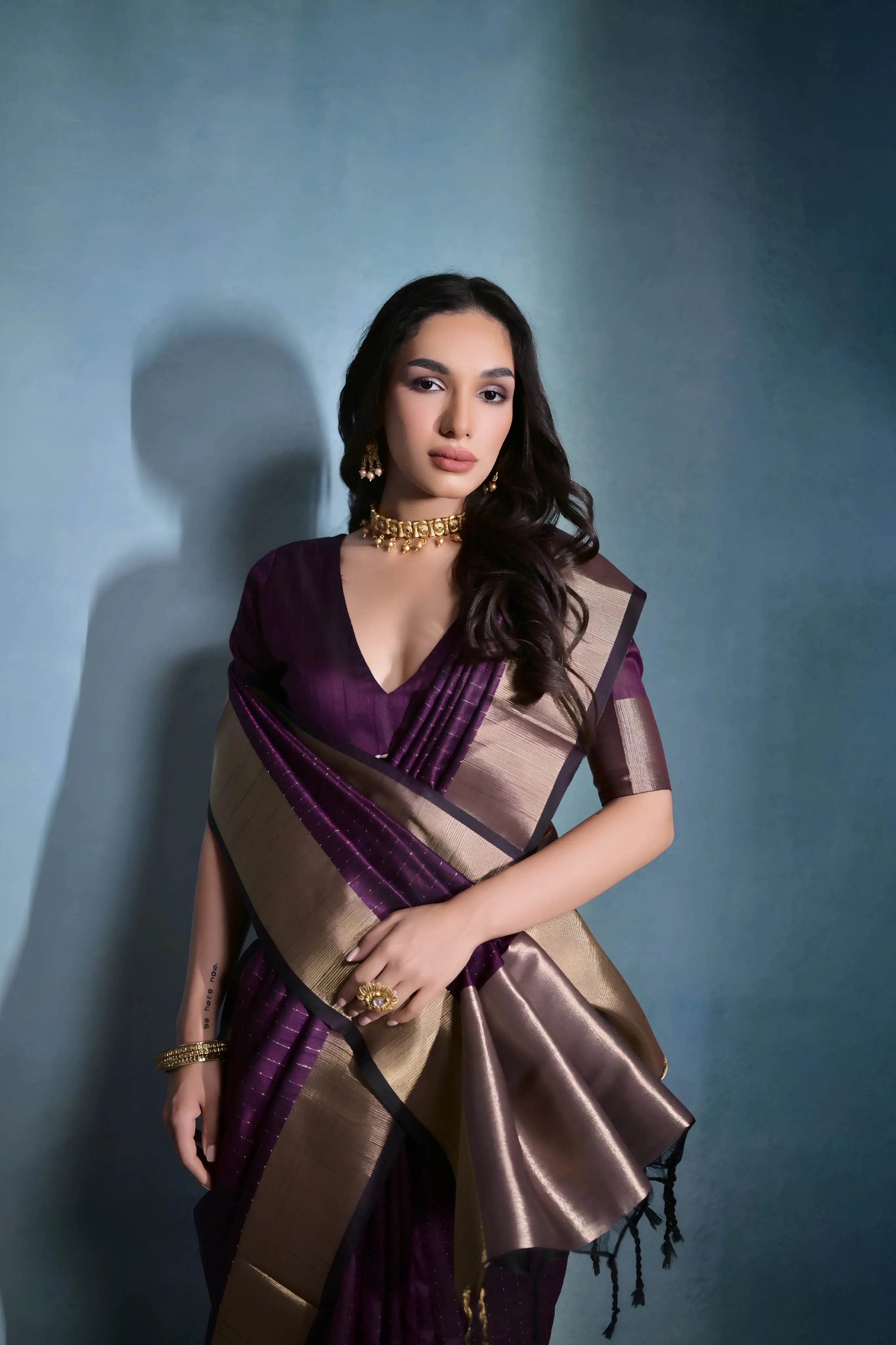 Woman wearing a purple silk saree with a gold border and traditional jewelry, standing against a blue background.