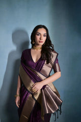 Woman wearing a purple silk saree with a gold border and traditional jewelry, standing against a blue background.