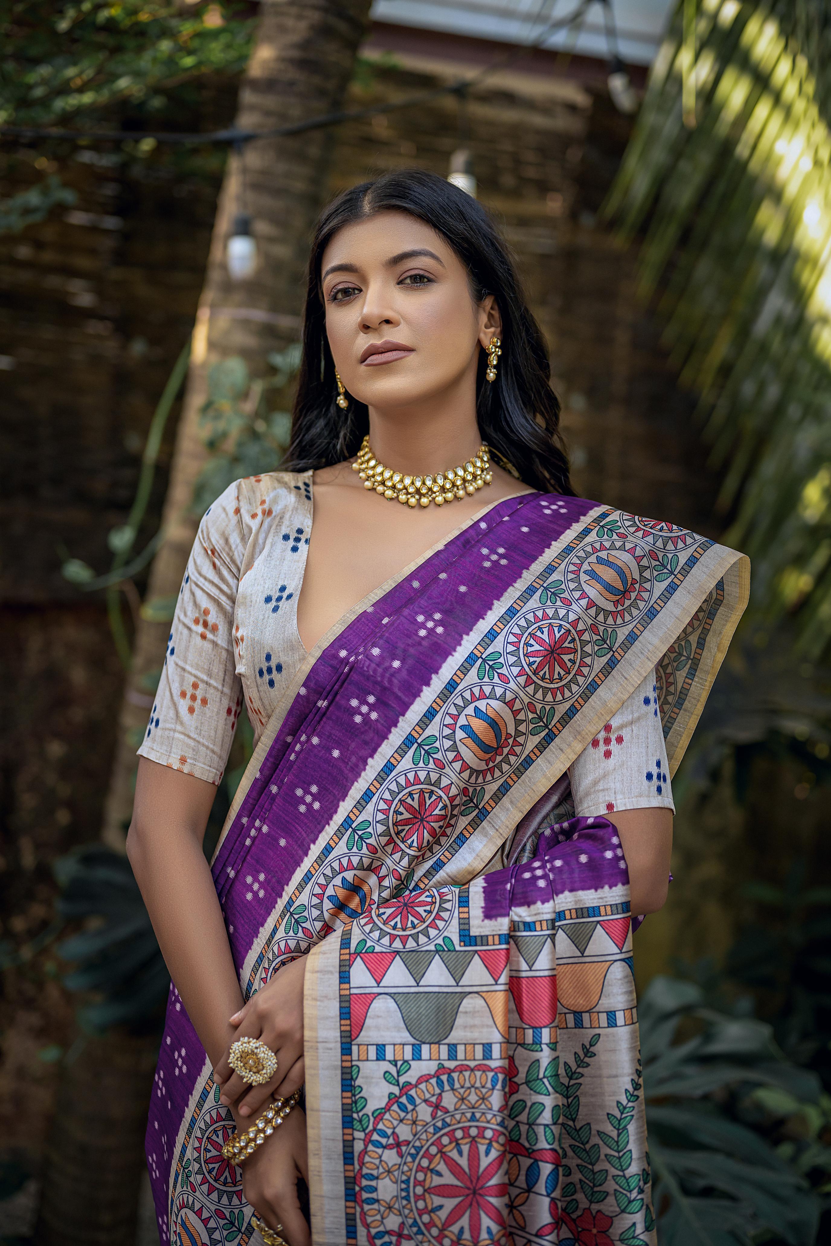 A woman models a Royal Purple Tussar Silk Saree with small white scattered prints. The pallu features a wide, elaborate panel with intricate Madhubani-style folk art prints in white, red, green, and blue. She wears a cream/gold embellished blouse and a gold Kundan choker necklace.