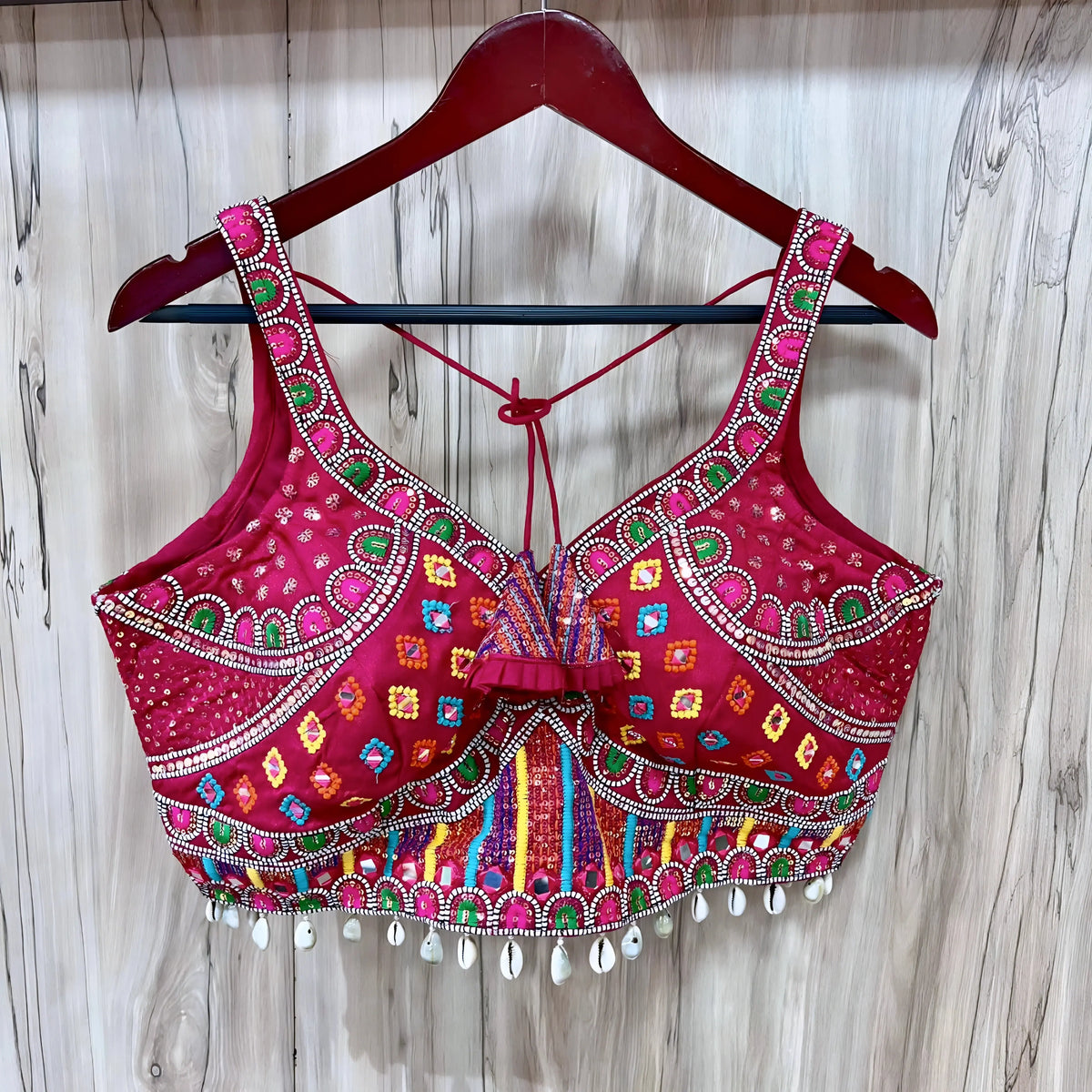 raditional rani pink Gujarati blouse featuring vibrant multicolor geometric embroidery in green, yellow, blue, and orange, adorned with intricate mirror work and delicate white cowrie shell trim along the hem. The design includes wide embroidered shoulder straps, structured cups, decorative front ruffle, and tasseled tie-back detail, displayed flat on a light wood panel background.