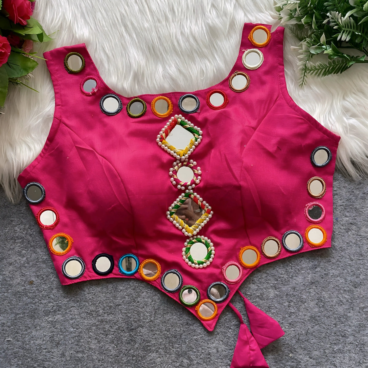  Rani pink ethnic blouse featuring vibrant mirror work with multicolored embroidered borders and decorative diamond and round mirrors arranged vertically at the center, accented with pearl detailing and a tie-up tassel at the waist. Displayed flat on a grey surface with a white faux fur background and green leafy accents, highlighting its festive and traditional design.