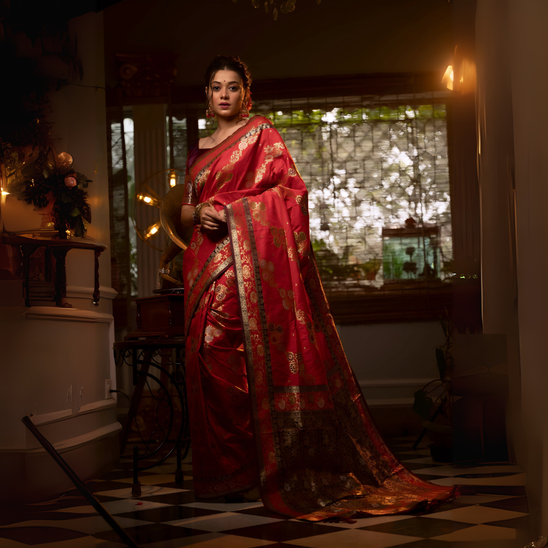 A woman models a luxurious Deep Red Soft Banarasi Silk Saree. The saree features a wide border and pallu with dense, intricate gold Zari weaving in traditional floral and paisley patterns. She wears a dark contrasting blouse and large gold jewelry, posing indoors on a black and white checkered floor.