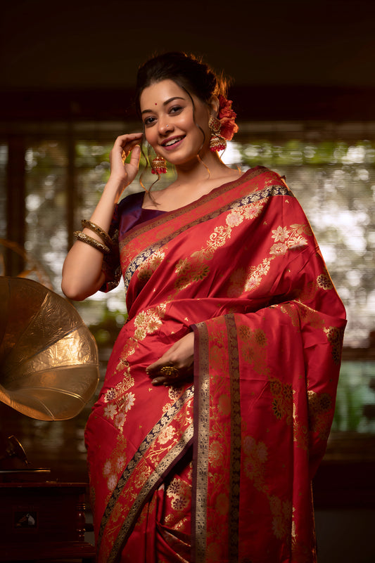 A woman models a luxurious Deep Red Soft Banarasi Silk Saree. The saree features a wide border and pallu with dense, intricate gold Zari weaving in traditional floral and paisley patterns. She wears a dark contrasting blouse and large gold jewelry, posing indoors on a black and white checkered floor.