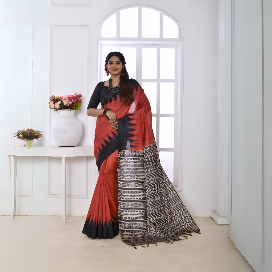 A woman models a striking Terracotta Red Soft Tussar Silk Saree. The saree features a bold black zigzag pattern along the border and a pallu with dense geometric black and white tribal-style print. She wears a black blouse and chunky oxidized silver jewelry, posing indoors by a large window.