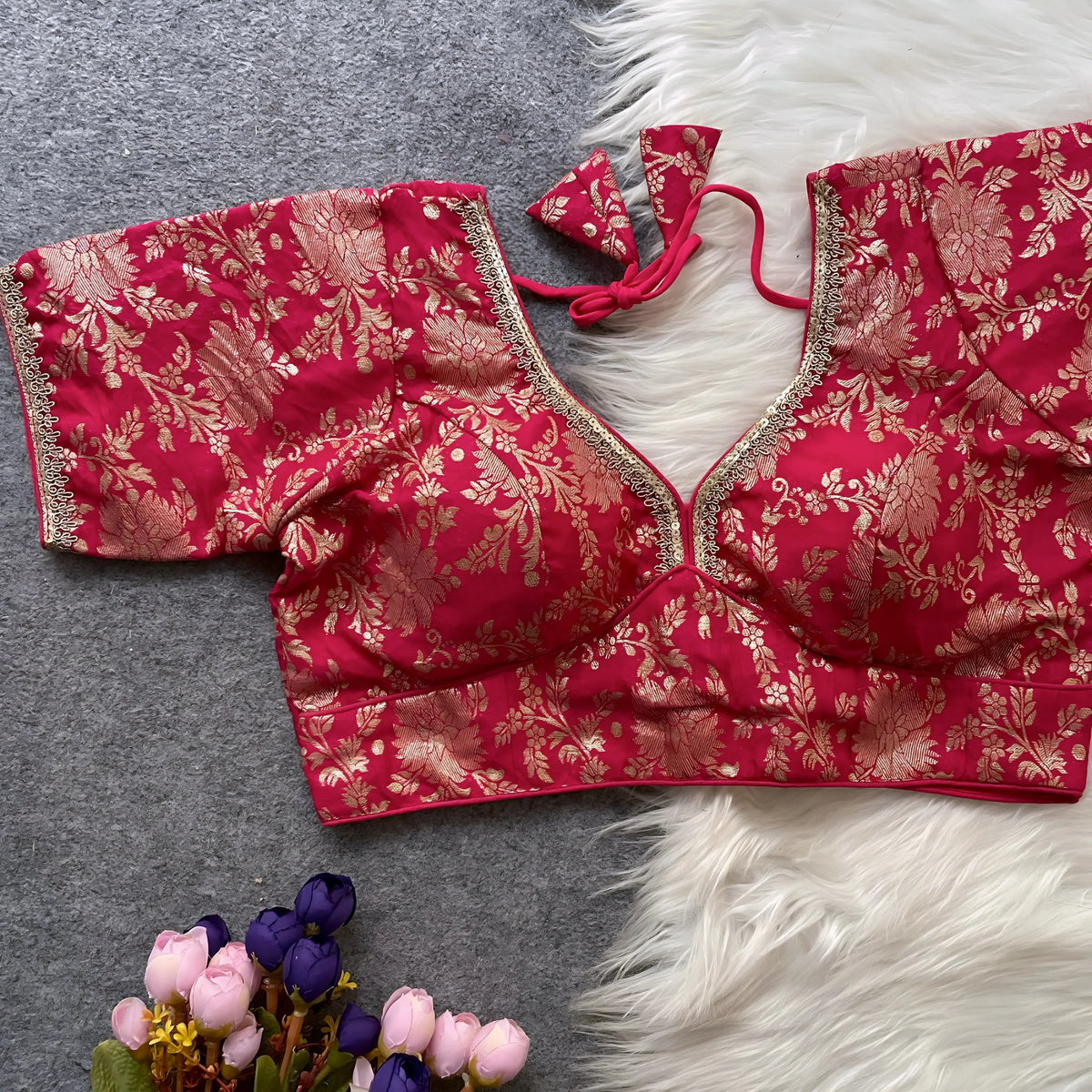 Red brocade blouse featuring intricate gold floral zari work and decorative trim along the neckline and sleeves, displayed flat on a grey surface with a white faux fur accent and a bunch of artificial pink and purple flowers, capturing its festive ethnic design and elegant craftsmanship.