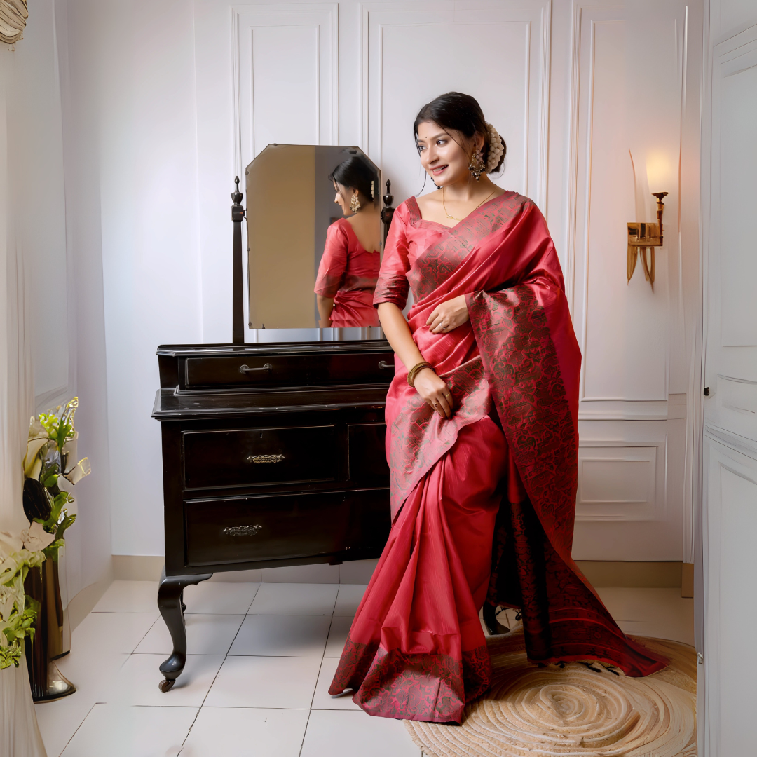 A woman models a luxurious Coral Red Raw Silk Saree. The saree features a wide border and pallu with dense, intricate weaving in a contrasting dark brown/black Kalamkari-style pattern. She wears a matching short-sleeve red blouse and traditional gold jewelry, with white flowers adorning her hair, posing indoors next to a dark wooden vanity.