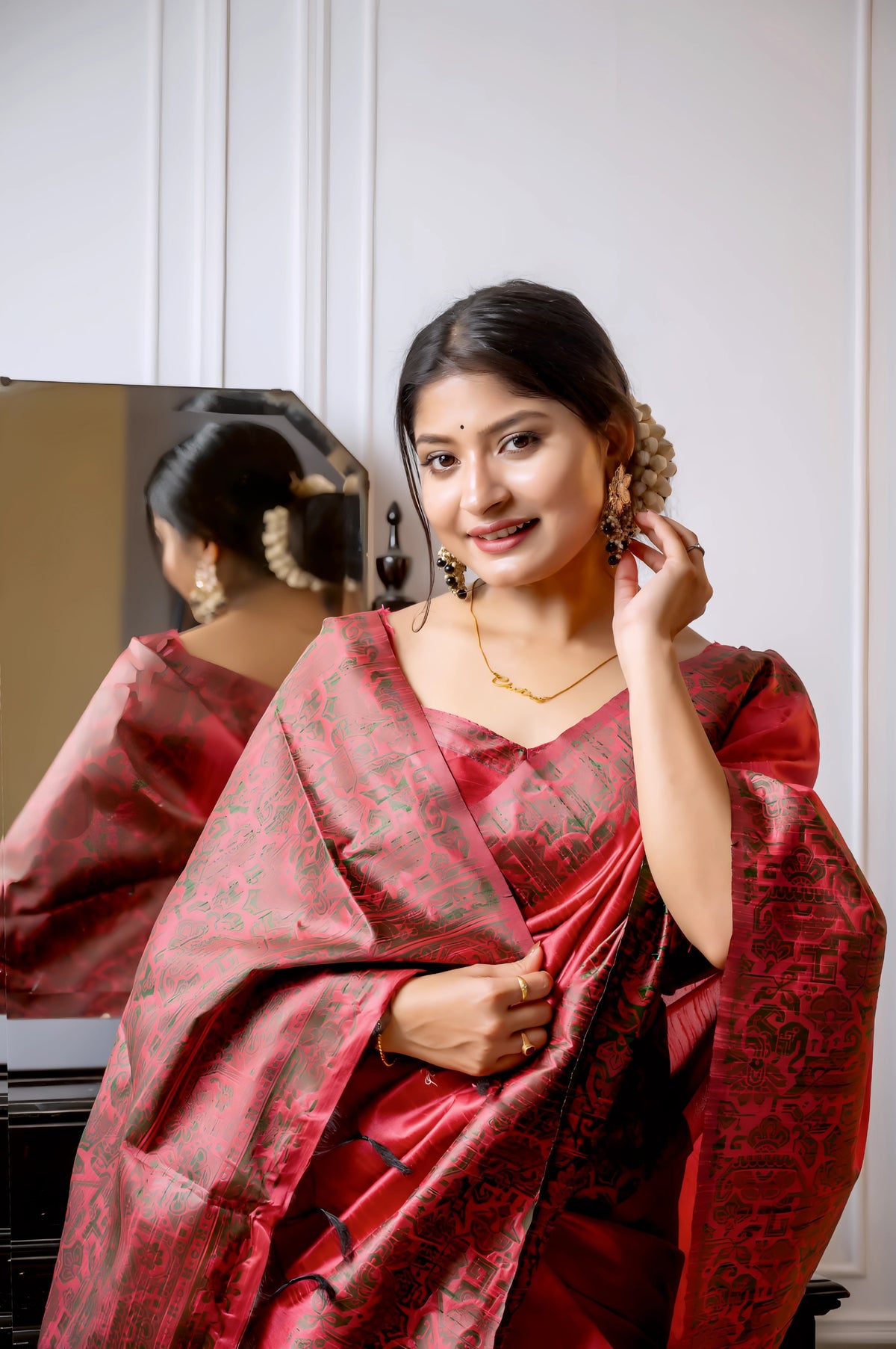A woman models a luxurious Coral Red Raw Silk Saree. The saree features a wide border and pallu with dense, intricate weaving in a contrasting dark brown/black Kalamkari-style pattern. She wears a matching short-sleeve red blouse and traditional gold jewelry, with white flowers adorning her hair, posing indoors next to a dark wooden vanity.