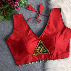Red padded blouse with green piping, featuring a central embroidered triangle motif with mirror work and shell detailing along the hem, displayed on a grey surface with artificial pink flowers and white fur in the background.