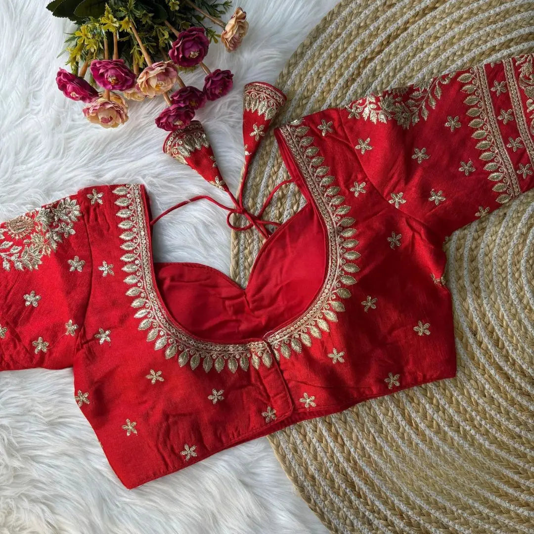  Red embroidered silk blouse with intricate gold zari floral patterns and tie-back detail, displayed on a white fur background with a bunch of pink and peach flowers in the corner