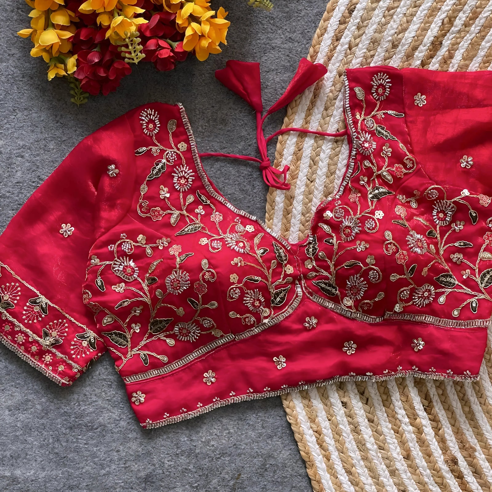 Red padded blouse with intricate gold floral embroidery and sequin accents, featuring a tie-back detail, displayed on a grey surface with artificial yellow and red flowers and a woven mat in the background.