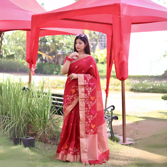A woman models a vibrant Deep Red Linen Silk Saree with delicate gold Zari weaving depicting colorful bird and floral motifs. The saree has a wide metallic gold border. She wears a sleeveless red blouse and striking red and white bangles, posing outdoors under a red canopy.