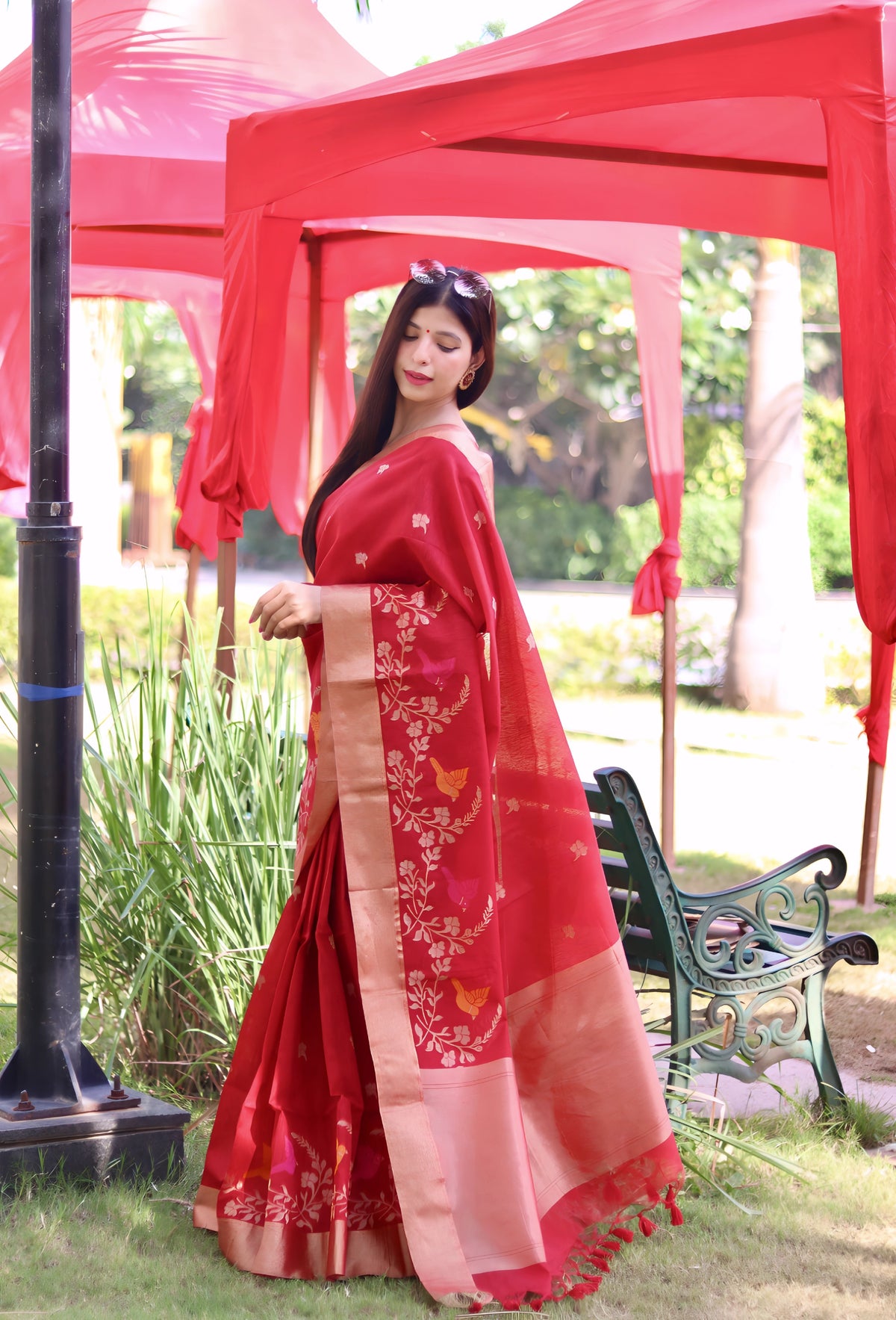 A woman models a vibrant Deep Red Linen Silk Saree with delicate gold Zari weaving depicting colorful bird and floral motifs. The saree has a wide metallic gold border. She wears a sleeveless red blouse and striking red and white bangles, posing outdoors under a red canopy.