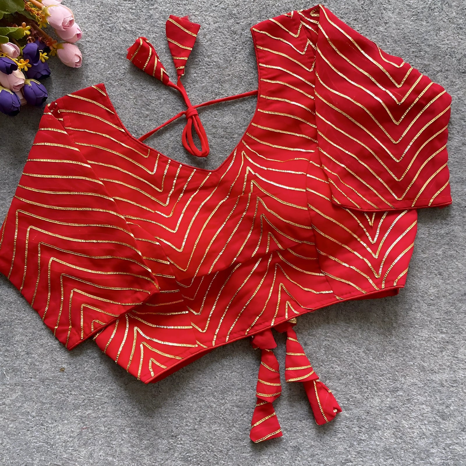  Red blouse adorned with gold gota patti zigzag embroidery, three-quarter sleeves, and a stylish back tie-up with coordinated triangular tassels. Displayed flat on a grey textured surface beside fresh pink and purple flowers, showcasing its festive and elegant ethnic style.