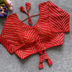  Red blouse adorned with gold gota patti zigzag embroidery, three-quarter sleeves, and a stylish back tie-up with coordinated triangular tassels. Displayed flat on a grey textured surface beside fresh pink and purple flowers, showcasing its festive and elegant ethnic style.
