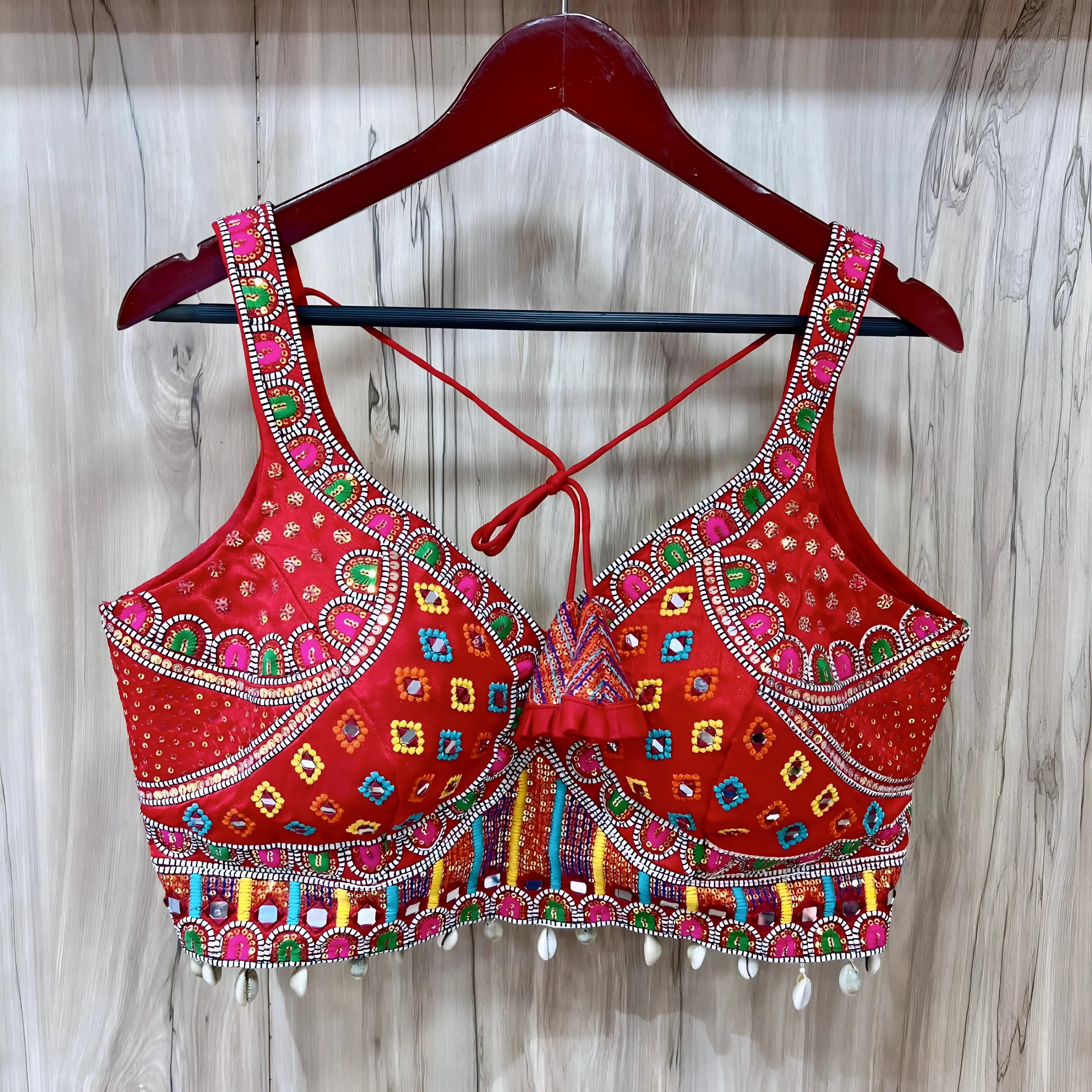 Traditional red Gujarati blouse featuring vibrant multicolor geometric and floral embroidery accented with mirror work and white cowrie shell trim along the hem. The blouse has wide embroidered straps, structured cups, a decorative front ruffle, and tasseled tie-back detail, displayed on a wooden hanger against a light wood panel background.