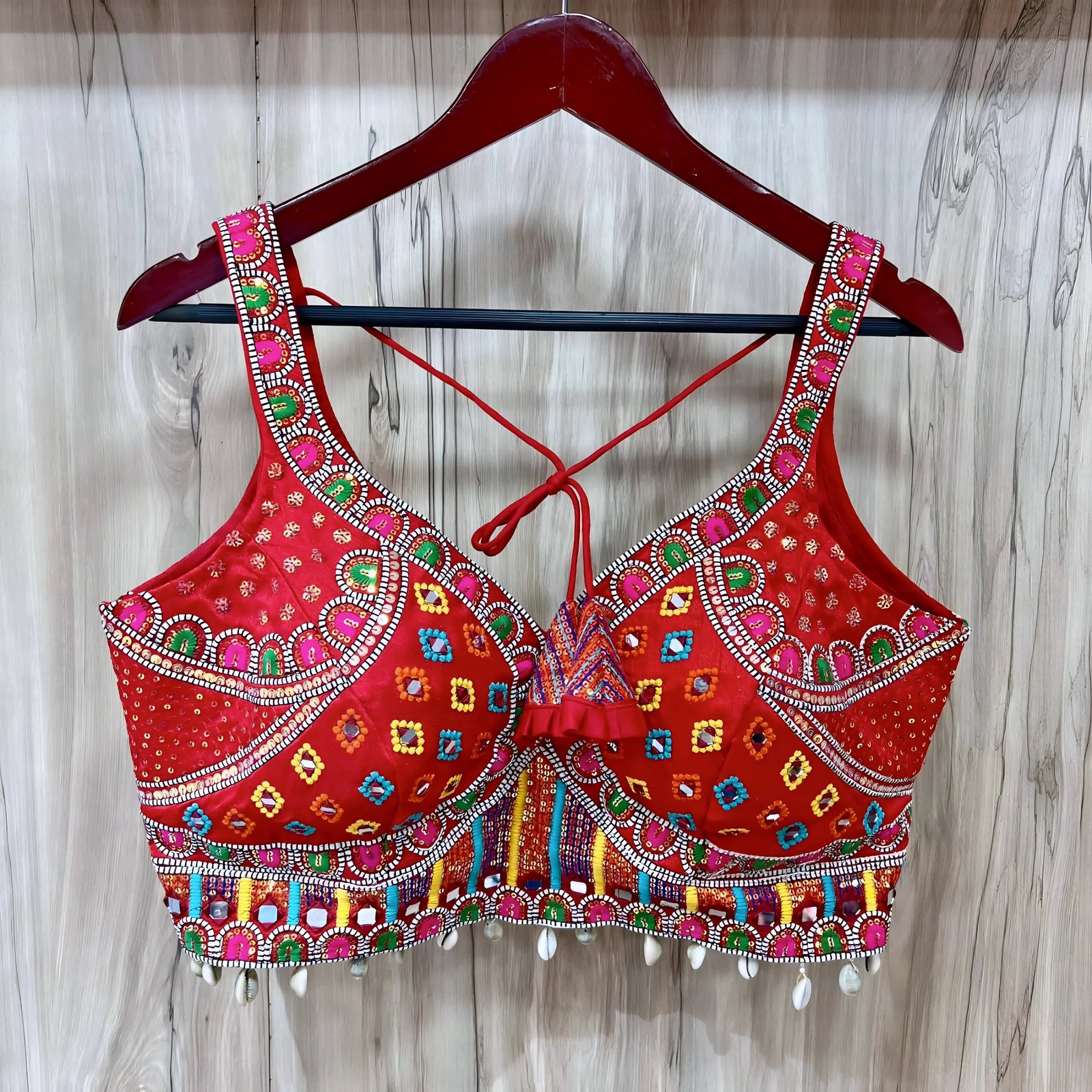 Traditional red Gujarati blouse featuring vibrant multicolor geometric and floral embroidery accented with mirror work and white cowrie shell trim along the hem. The blouse has wide embroidered straps, structured cups, a decorative front ruffle, and tasseled tie-back detail, displayed on a wooden hanger against a light wood panel background.