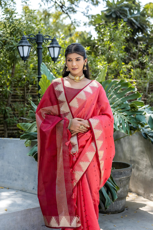 A woman models a striking Coral Red Tussar Silk Saree with scattered dark motifs. The saree has a wide border and pallu featuring traditional gold Zari weaving interspersed with rich maroon panels. She wears a gold choker necklace and is posed in a garden setting.