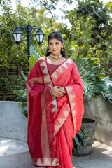 A woman models a striking Coral Red Tussar Silk Saree with scattered dark motifs. The saree has a wide border and pallu featuring traditional gold Zari weaving interspersed with rich maroon panels. She wears a gold choker necklace and is posed in a garden setting.