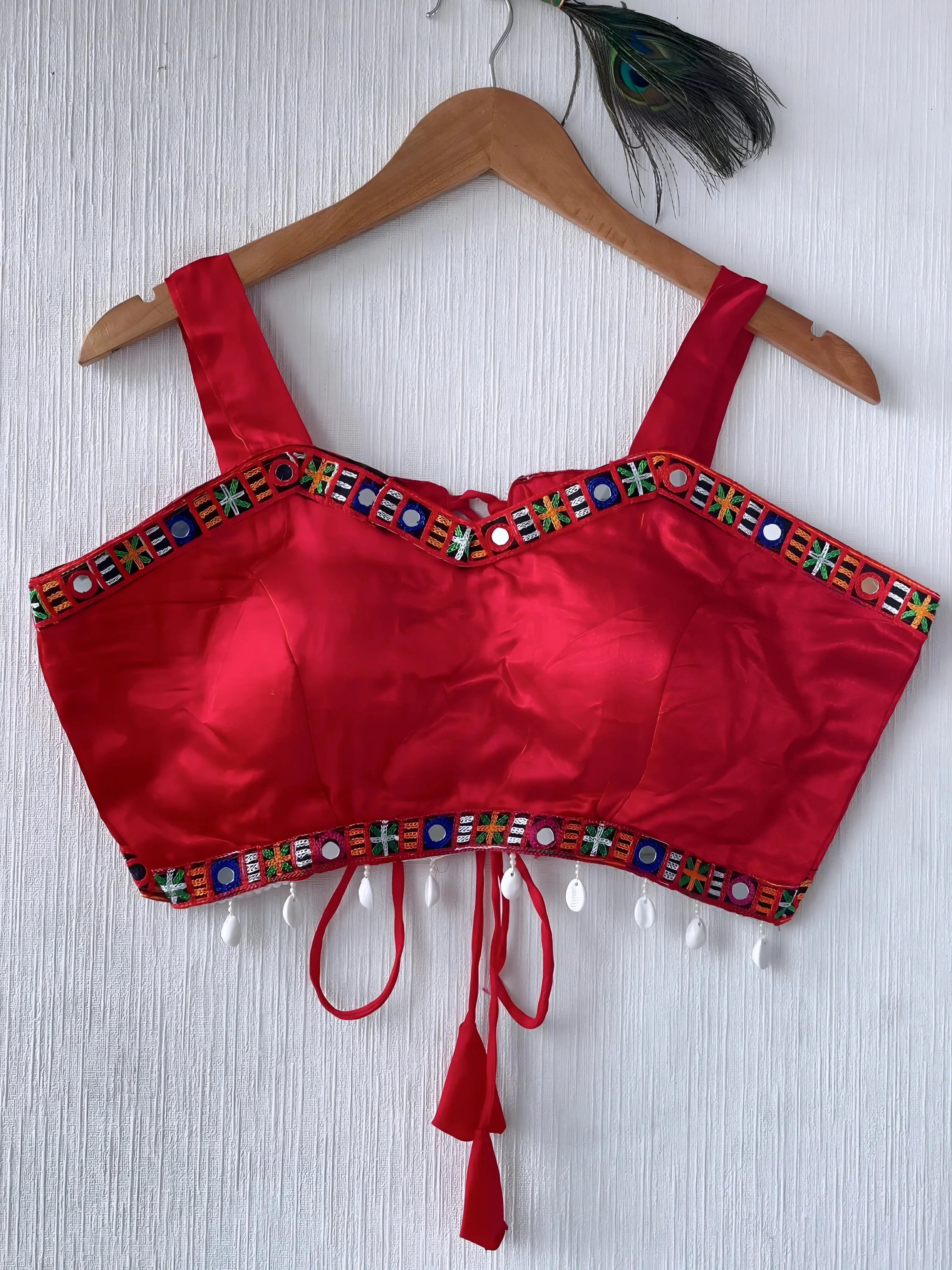  Bright red ethnic blouse featuring colorful mirror work and multicolored embroidered geometric border in green, blue, and orange, adorned with white shell tassels and a tie-up waist detail. Displayed on a wooden hanger against a textured off-white wall with peacock feathers, showcasing a festive and traditional look.