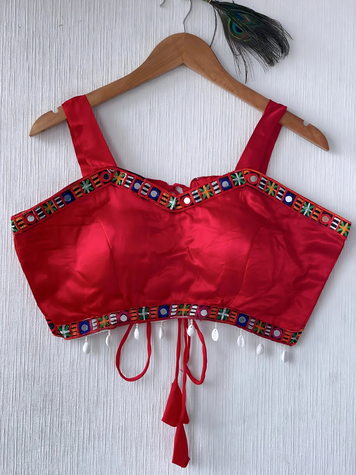 Bright red ethnic blouse featuring colorful mirror work and multicolored embroidered geometric border in green, blue, and orange, adorned with white shell tassels and a tie-up waist detail. Displayed on a wooden hanger against a textured off-white wall with peacock feathers, showcasing a festive and traditional look.