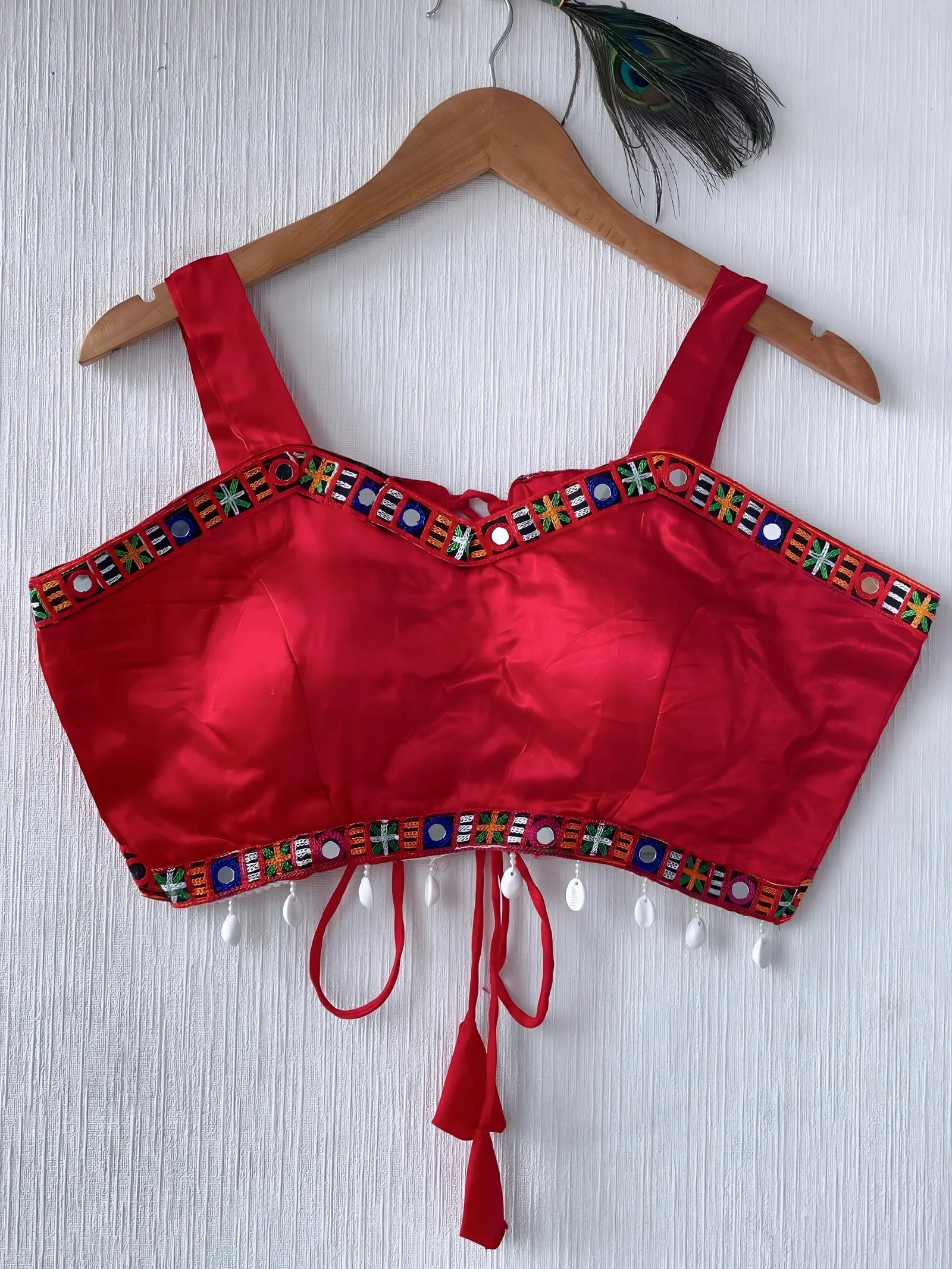  Bright red ethnic blouse featuring colorful mirror work and multicolored embroidered geometric border in green, blue, and orange, adorned with white shell tassels and a tie-up waist detail. Displayed on a wooden hanger against a textured off-white wall with peacock feathers, showcasing a festive and traditional look.