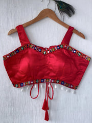  Bright red ethnic blouse featuring colorful mirror work and multicolored embroidered geometric border in green, blue, and orange, adorned with white shell tassels and a tie-up waist detail. Displayed on a wooden hanger against a textured off-white wall with peacock feathers, showcasing a festive and traditional look.