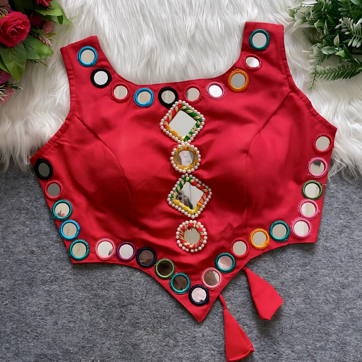 Red ethnic blouse featuring intricate mirror work with multicolored embroidered borders and embellished diamond-shaped mirrors at the center, finished with decorative tassel tie-ups at the waist. Displayed flat on a grey surface with white faux fur and floral accents for a festive and traditional look.