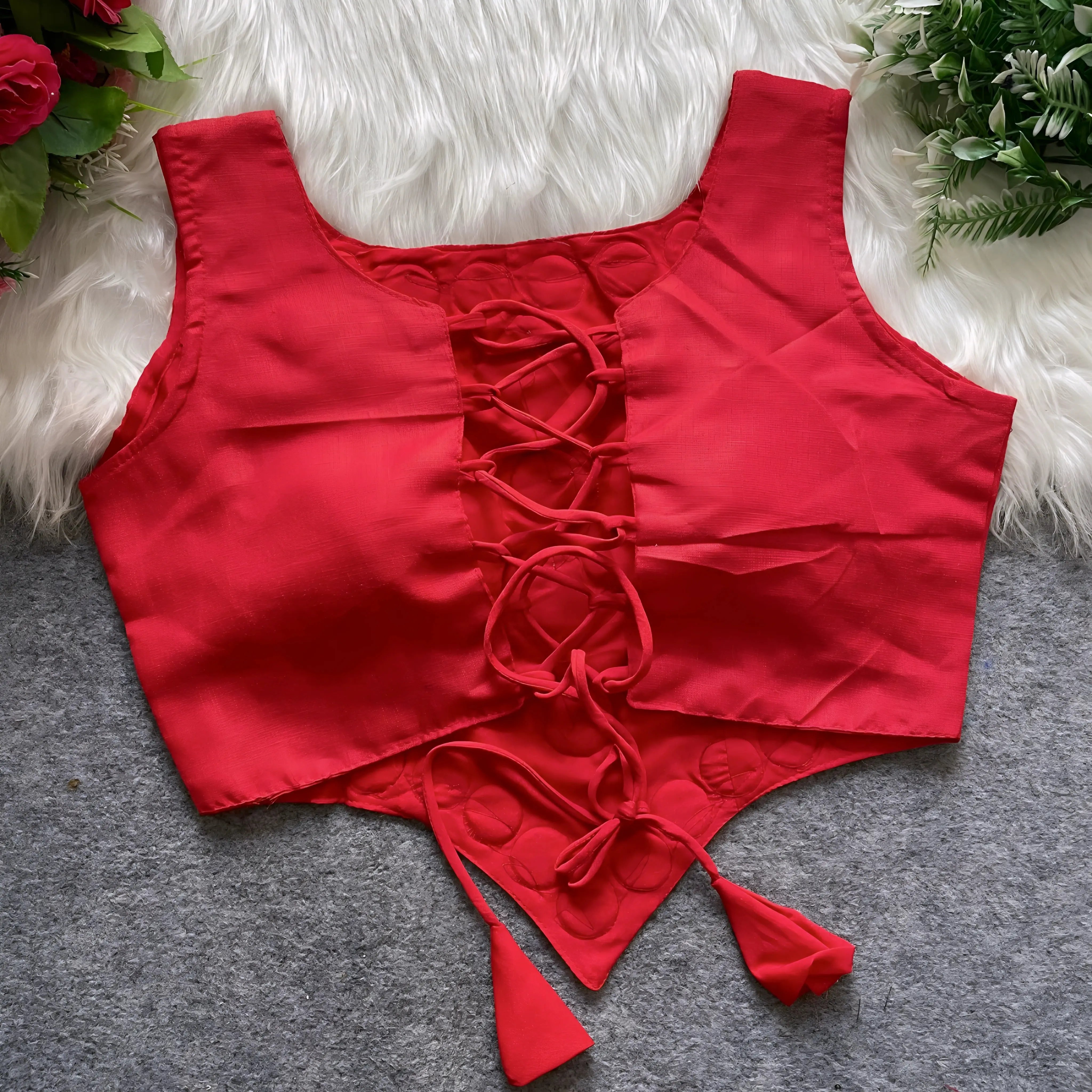 Red ethnic blouse featuring intricate mirror work with multicolored embroidered borders and embellished diamond-shaped mirrors at the center, finished with decorative tassel tie-ups at the waist. Displayed flat on a grey surface with white faux fur and floral accents for a festive and traditional look.