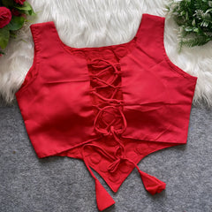 Red ethnic blouse featuring intricate mirror work with multicolored embroidered borders and embellished diamond-shaped mirrors at the center, finished with decorative tassel tie-ups at the waist. Displayed flat on a grey surface with white faux fur and floral accents for a festive and traditional look.