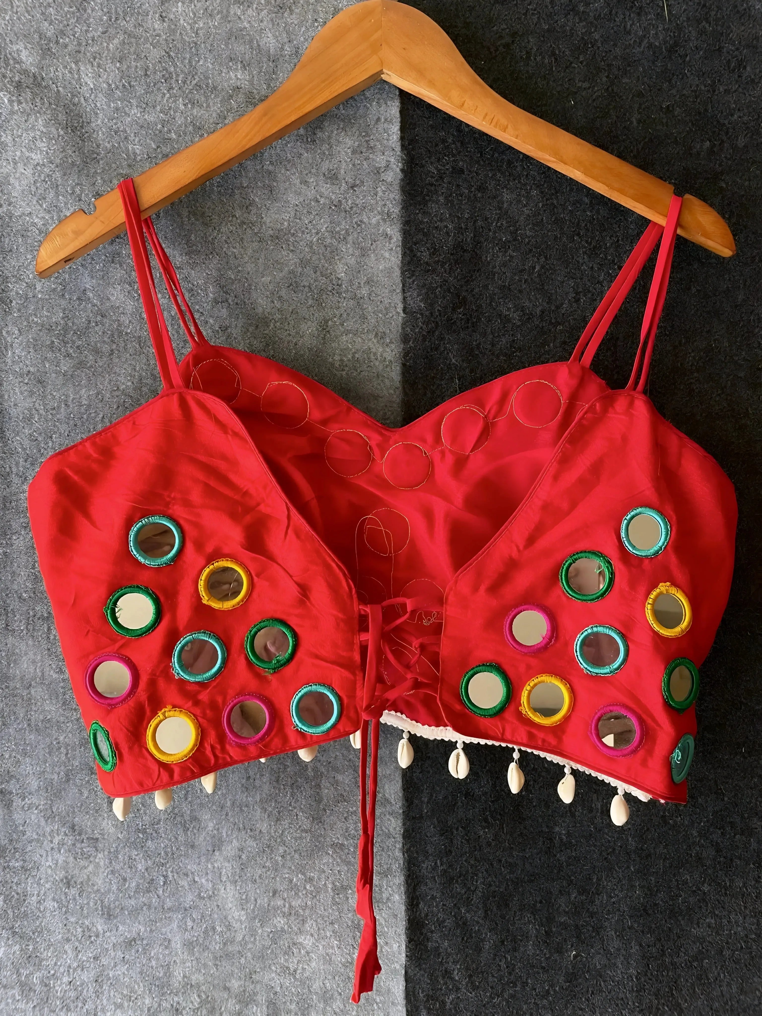 Red corset-style blouse decorated with vibrant round mirrors and multicolored embroidered borders, featuring thin shoulder straps and shell tassel drops along the hem. Displayed on a wooden hanger against a two-tone grey textured background, highlighting its festive and modern ethnic appeal.
