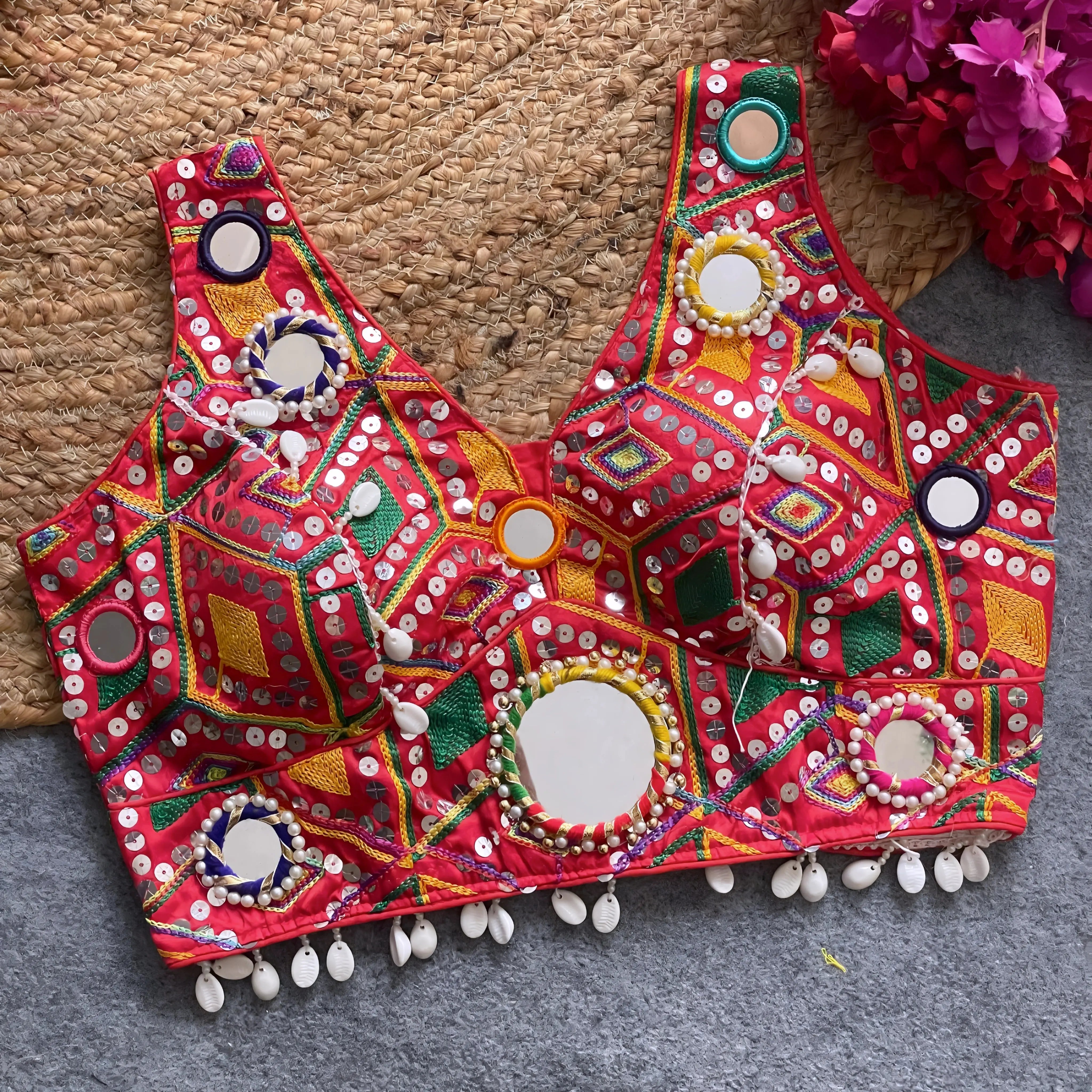 Traditional red Gujarati-style blouse richly adorned with vibrant multicolor geometric embroidery, large circular mirror work bordered by colorful bead rings, white cowrie shell trim along the hem, and decorative tassels. The blouse is displayed flat on a grey carpet beside a woven mat and vibrant fuchsia artificial flowers.