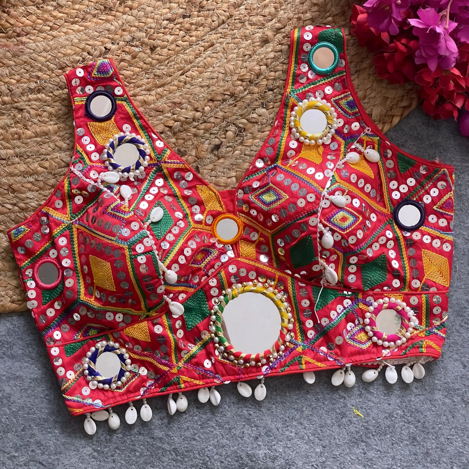Traditional red Gujarati-style blouse richly adorned with vibrant multicolor geometric embroidery, large circular mirror work bordered by colorful bead rings, white cowrie shell trim along the hem, and decorative tassels. The blouse is displayed flat on a grey carpet beside a woven mat and vibrant fuchsia artificial flowers.