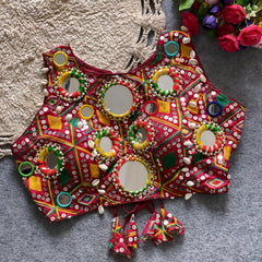 Traditional red Kutch-style blouse adorned with vibrant multicolor geometric embroidery, large circular mirrors bordered by colorful bead rings, cowrie shell accents, and tasseled tie-back detail, displayed flat on a grey carpet beside a cream woven mat and a bouquet of artificial roses.
