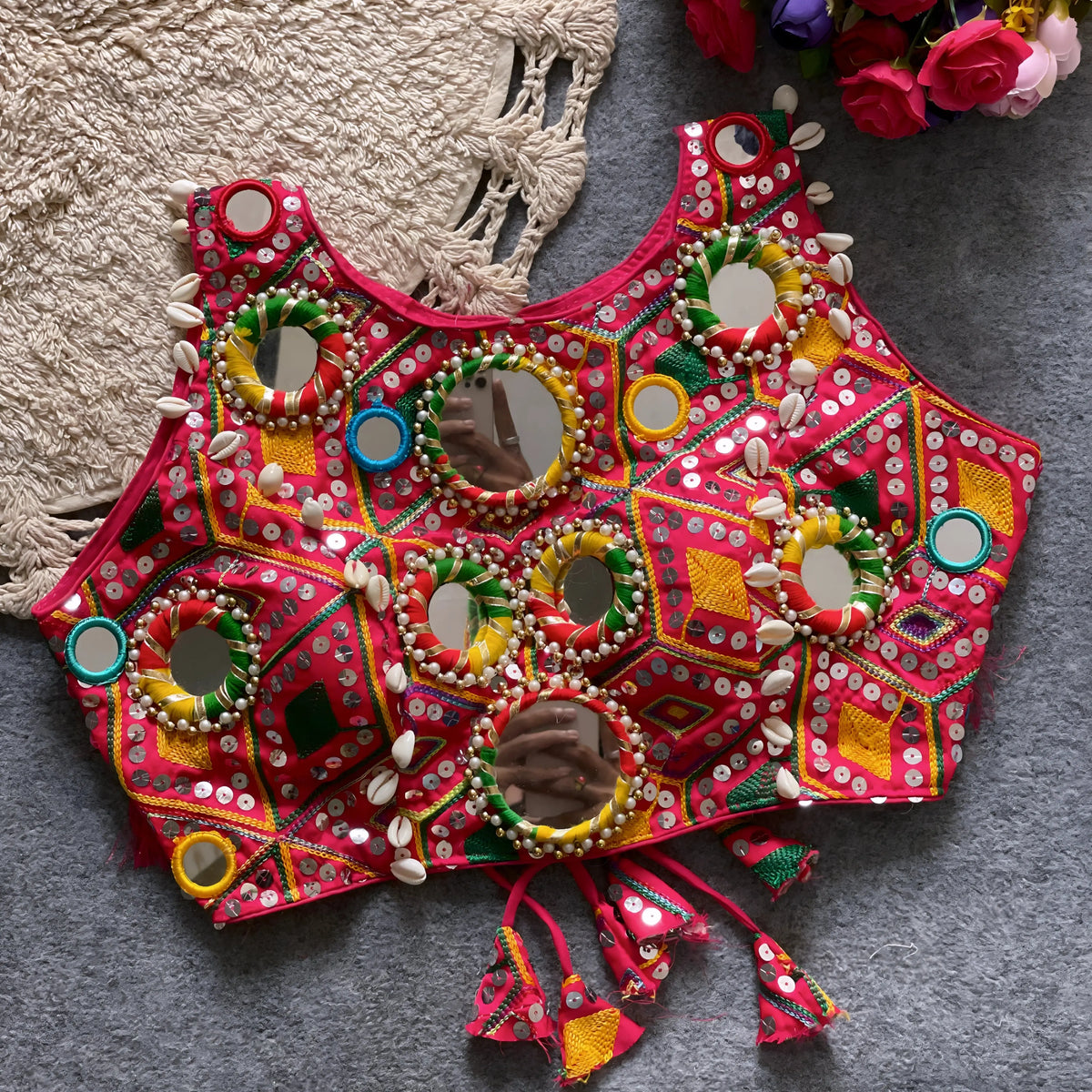 raditional red Kutch-style blouse adorned with vibrant multicolor geometric embroidery, large circular mirrors bordered by colorful bead rings, cowrie shell accents, and tasseled tie-back detail, displayed flat on a grey carpet beside a cream woven mat and a bouquet of artificial roses.